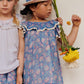 Indoors, two children stand with serious expressions; one wears a Louise Misha Girls' Natiya Dress holding flowers, the other is in a white top and shorts.