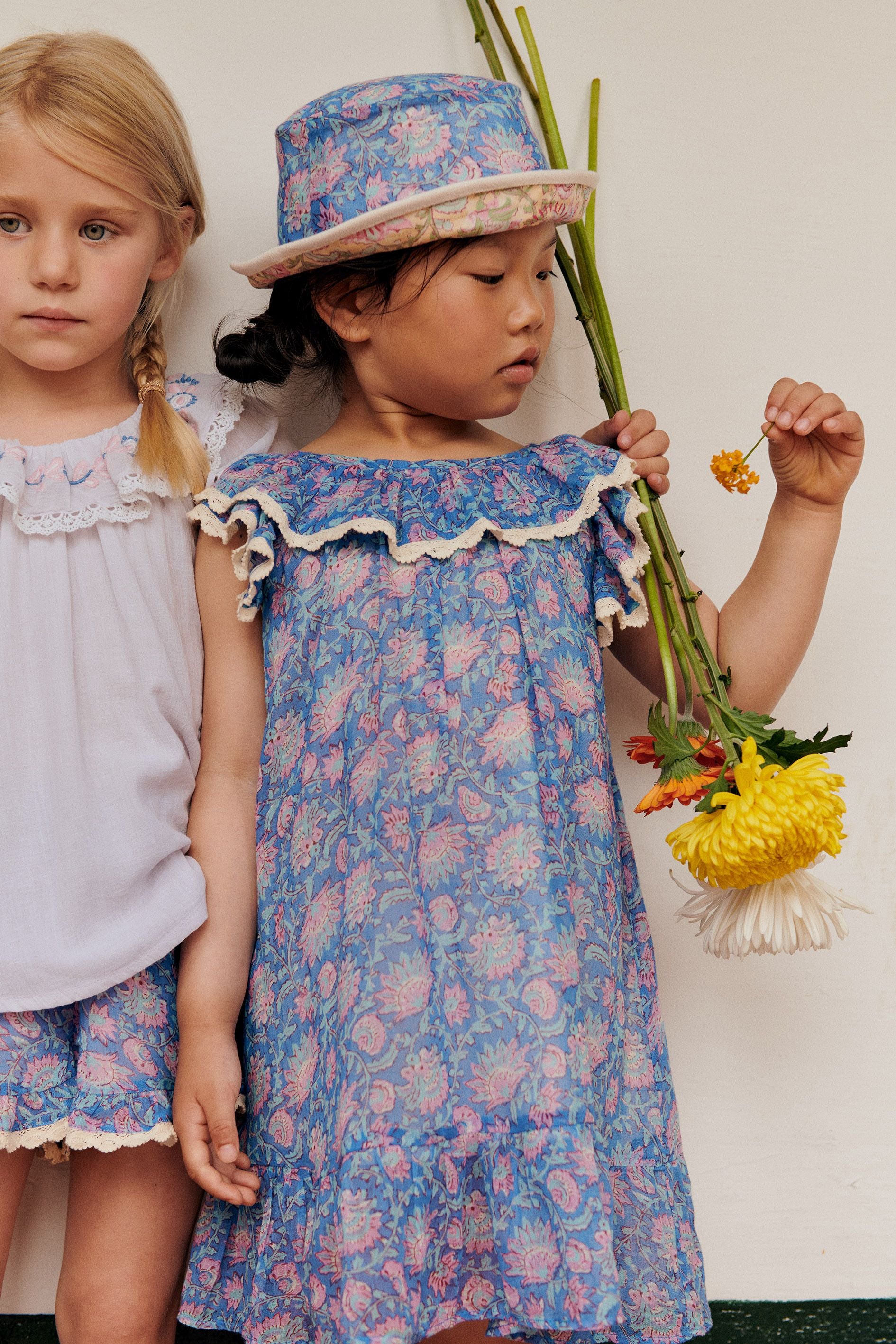 Indoors, two children stand with serious expressions; one wears a Louise Misha Girls' Natiya Dress holding flowers, the other is in a white top and shorts.