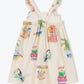 The Girls' Cocos Frios Dress by Arsene & Les Pipelettes is a sleeveless cream cotton twill dress with ruffled straps, adorned with vibrant tropical prints of toucans, palm trees, fruit stands, and "Hay cocos frios" signs—perfect for a playful look.