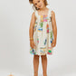 A young girl stands barefoot on a white background, wearing the vibrant Girls' Cocos Frios Dress by Arsene & Les Pipelettes, crafted from soft cotton twill with colorful prints and delicate ruffled straps. A coconut and two oranges rest on the floor beside her.