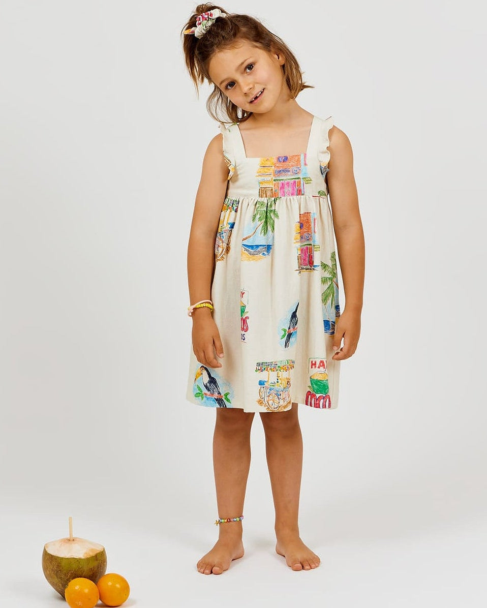 A young girl stands barefoot on a white background, wearing the vibrant Girls' Cocos Frios Dress by Arsene & Les Pipelettes, crafted from soft cotton twill with colorful prints and delicate ruffled straps. A coconut and two oranges rest on the floor beside her.
