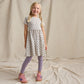 A smiling young girl with blonde braided hair wears the Coop Kids PC Girls’ Sunny Dress made from organic Pima cotton, paired with lavender leggings and pink sneakers, standing against a beige fabric backdrop.