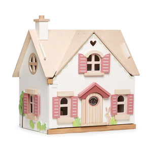 A Tenderleaf Cottontail Doll Cottage by Tender Leaf Toys with a chimney, pink shutters, and heart-shaped window cutouts is depicted. The front features a small porch and decorative plants on either side. The self-assembly kit offers nostalgic charm reminiscent of a quaint countryside cottage.
