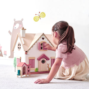 A young girl plays with a Tender Leaf Toys Tenderleaf Cottontail Doll Cottage, adjusting its window shutters. The backdrop features a wall decal of a bear and butterfly, evoking the charm of a countryside cottage with its retro style furniture.