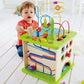 A young child in a red shirt is playing with the Hape Country Critters Play Cube, which features a vibrant maze, shape sorters, and various interactive elements, all on a light-colored floor.