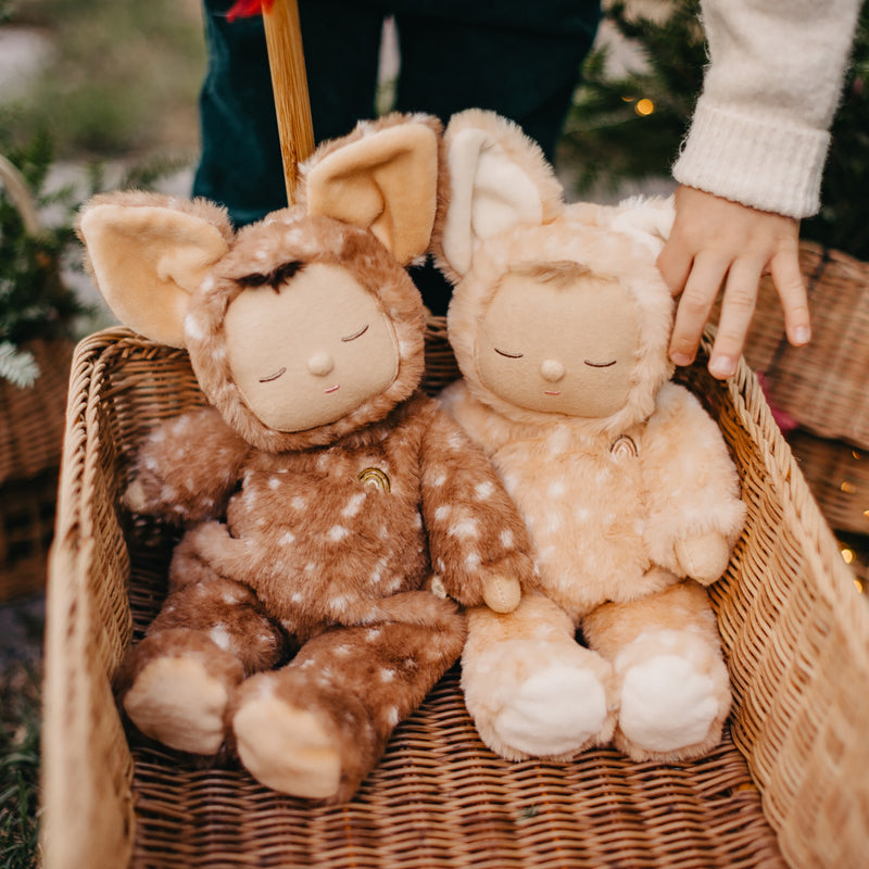 Two plush Olli Ella Cozy Dinkum Dolls dressed in animal costumes with closed eyes sit in a wicker basket, while a child's hand rests on one toy. These posable dolls make for the perfect newborn gift.