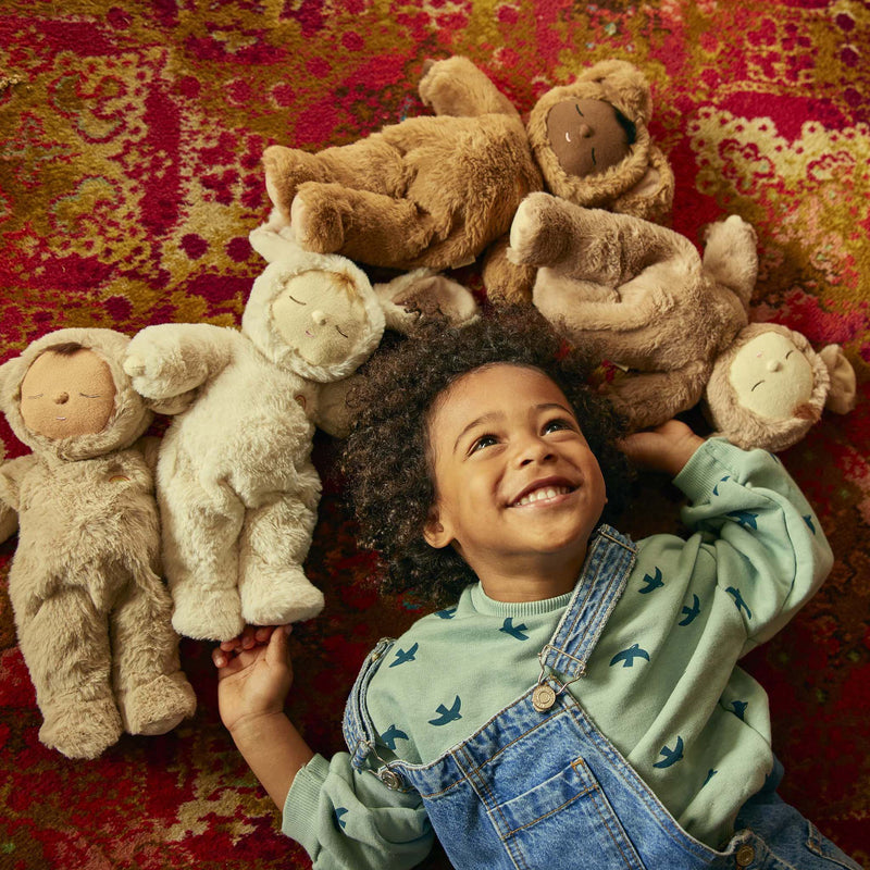 A young child in a green shirt and denim overalls lies on a colorful rug, smiling, surrounded by cozy Olli Ella Cozy Dinkum Dolls and other stuffed animal toys.