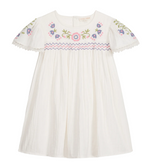 The Girls’ Ana Dress by Louise Misha is a white cotton dress with short sleeves, adorned with colorful floral embroidery on the chest and sleeves, hand-smocked details along the embroidered yoke, pink and blue zigzag trim, and finished with delicate lace edging on the sleeves.