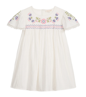 The Girls’ Ana Dress by Louise Misha is a white cotton dress with short sleeves, adorned with colorful floral embroidery on the chest and sleeves, hand-smocked details along the embroidered yoke, pink and blue zigzag trim, and finished with delicate lace edging on the sleeves.