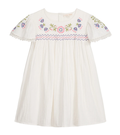 The Girls’ Ana Dress by Louise Misha is a white cotton dress with short sleeves, adorned with colorful floral embroidery on the chest and sleeves, hand-smocked details along the embroidered yoke, pink and blue zigzag trim, and finished with delicate lace edging on the sleeves.