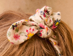 A close-up of the LM Chloe Scrunchie by Louise Misha, a light-colored floral hair accessory tied in brown hair.