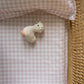 A small, knitted toy animal rests on a pink and white checkered blanket in a wicker crib with a Gooselings Picnic Gingham Pink Crib Sheet that's OEKO-TEX certified.