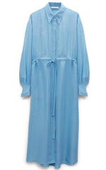 The Heritage Ease Dress by Dorothee Schumacher is a long, light blue washed silk shirtdress with a collar, long sleeves, concealed button placket, and drawstring waist, elegantly displayed against a white background.