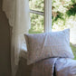 The Enchanted Wildflowers Toddler Pillow with Insert by Gooselings rests by a window with sheer curtains, overlooking a sunlit green garden, creating the ideal setting for nurturing dreams as nighttime gently unfolds.