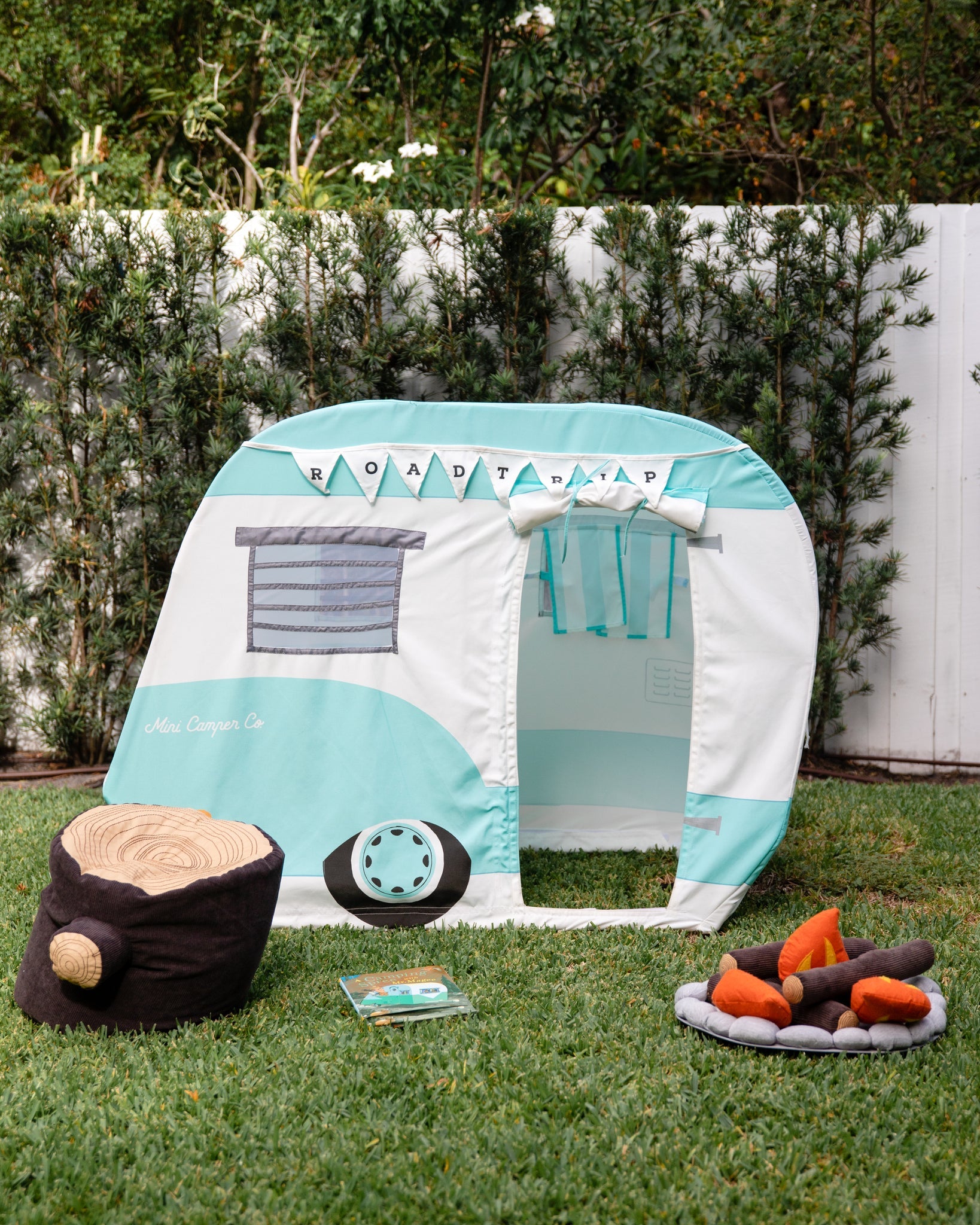 The Asweets children's play tent, designed to resemble a camper, is ideal for a playroom. It comes with comfortable pillows shaped like logs and the Asweets Tree Stump Poof, all accented by a backdrop of a white fence and vibrant greenery.