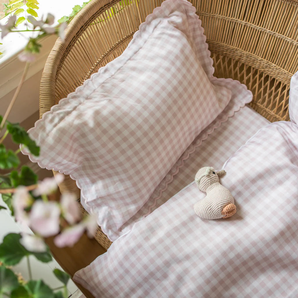 This charming baby gift features a Gooselings rattan crib with a Picnic Gingham Pink Toddler Duvet and Insert, scalloped pillows, and a knitted toy. A green plant and soft window light complete the serene setup.