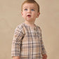 A toddler with light brown hair and blue eyes is wearing the Quincy Mae Baby Boys’ and Boys’ Rory Shirt, a beige and brown plaid long-sleeve top paired with matching pants, standing against a plain beige background.