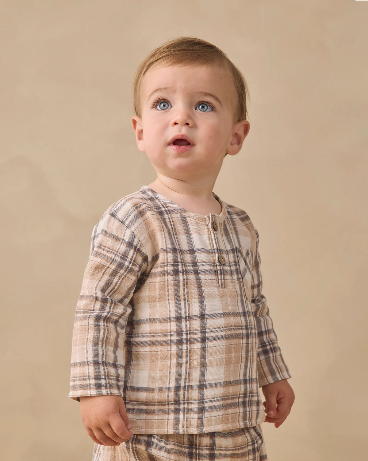 A toddler with light brown hair and blue eyes is wearing the Quincy Mae Baby Boys’ and Boys’ Rory Shirt, a beige and brown plaid long-sleeve top paired with matching pants, standing against a plain beige background.