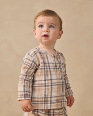 A toddler with light brown hair and blue eyes is wearing the Quincy Mae Baby Boys’ and Boys’ Rory Shirt, a beige and brown plaid long-sleeve top paired with matching pants, standing against a plain beige background.