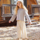 A young child with long blond hair stands outside a wooden cabin, wearing a knit hat, Rylee & Cru Girl’s Luna Long Sleeve, floral pants with a relaxed silhouette, and boots, looking upward and smiling for an effortless everyday look.