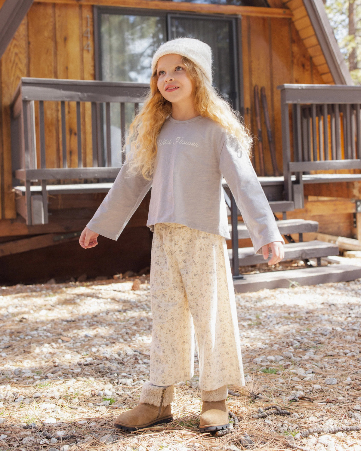 A young child with long blond hair stands outside a wooden cabin, wearing a knit hat, Rylee & Cru Girl’s Luna Long Sleeve, floral pants with a relaxed silhouette, and boots, looking upward and smiling for an effortless everyday look.