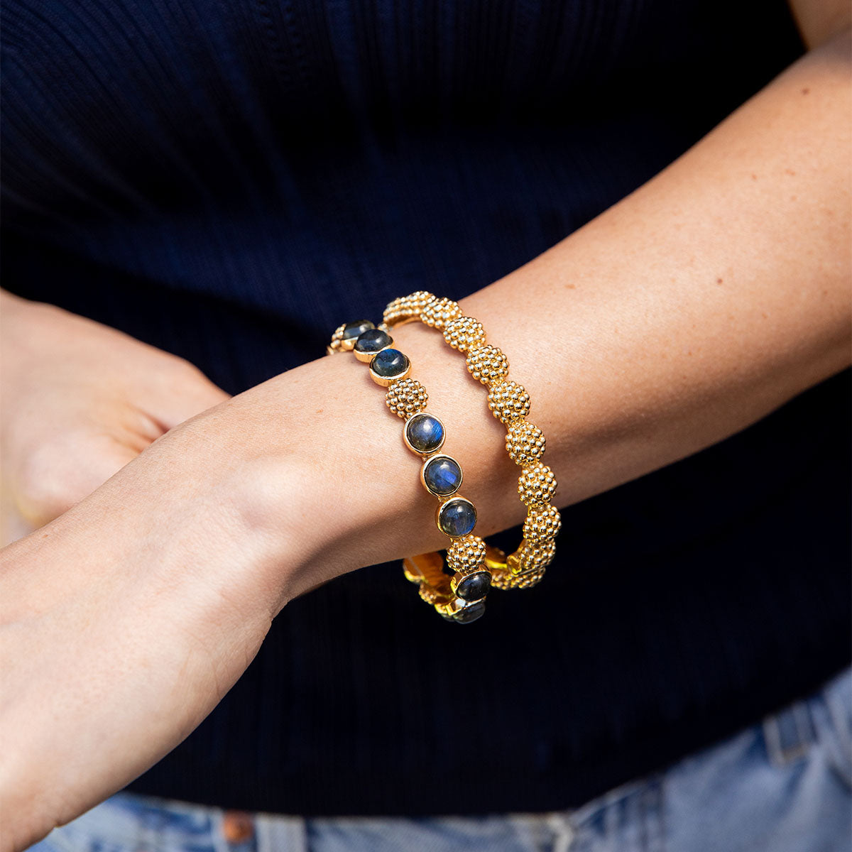 A person wearing two gold bracelets by Juliska Capucine De Wulf on their left wrist—including the Berry Gem Hinged Bangle—styled with a dark top and light jeans.