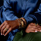 A person in a blue shirt and green pants showcases several stackable bracelets, including the Berry Small Hinged Bangle by Juliska Capucine De Wulf, along with golden bangles and rings on their hands.