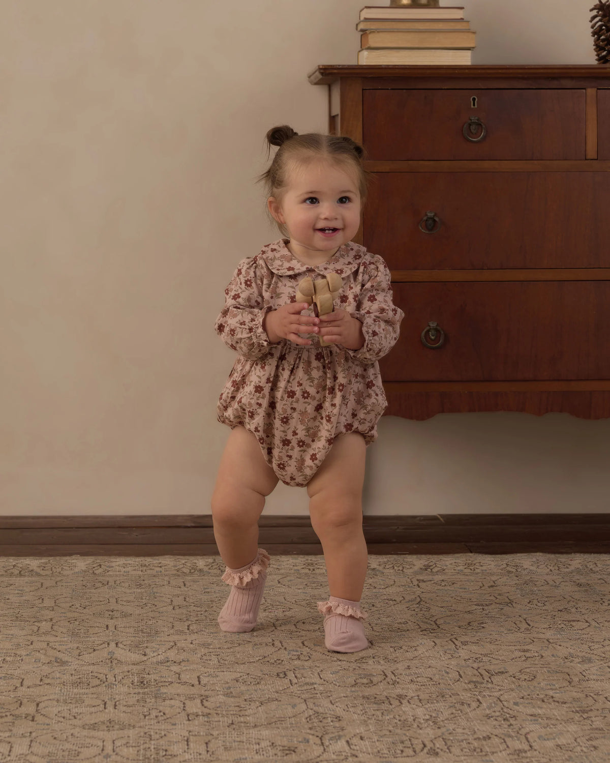 A toddler wearing the Rylee & Cru Baby Girls’ Cora Romper—with a charming floral print, a Peter Pan collar, and coconut buttons—stands on a carpeted floor holding a plush toy, in front of a wooden dresser topped with books.