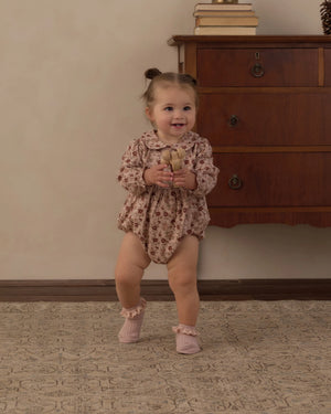 A toddler wearing the Rylee & Cru Baby Girls’ Cora Romper—with a charming floral print, a Peter Pan collar, and coconut buttons—stands on a carpeted floor holding a plush toy, in front of a wooden dresser topped with books.