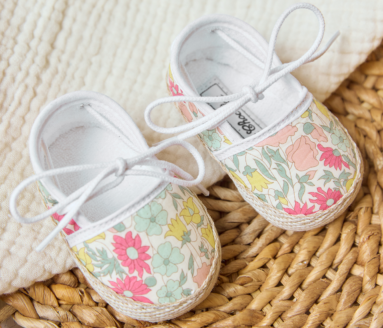 Baby Girls’ Liberty Fabric Espadrille Slipper by Tartine et Chocolat, featuring white laces and a pastel floral pattern, displayed on a textured woven mat beside cream-colored fabric.
