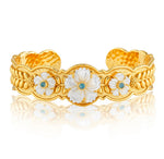 The Villandry Fleur Scalloped Cuff by Juliska Capucine De Wulf features a gold textured band adorned with three white floral motifs, each accented by a blue center, showcased against a white background.