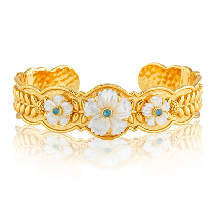 The Villandry Fleur Scalloped Cuff by Juliska Capucine De Wulf features a gold textured band adorned with three white floral motifs, each accented by a blue center, showcased against a white background.