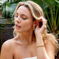 A woman with long blonde hair wears Juliska Capucine De Wulf Jasmine Hoop Earrings, along with a matching gold necklace, bracelet, and ring while posing outdoors among green plants.
