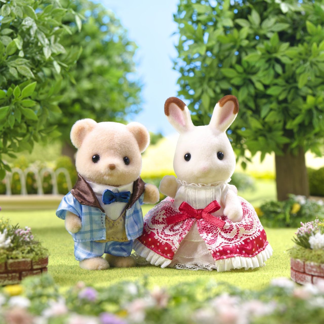 Two toy animal figures—Theo the bear in a blue suit and Freya Chocolate Rabbit in a red dress—from the Calico Critters 40th Anniversary Friends - Freya & Theo set stand side by side outdoors in a garden with trees and flowers, proudly showcasing their exclusive original outfits.