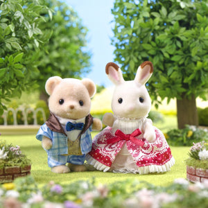 Two toy animal figures—Theo the bear in a blue suit and Freya Chocolate Rabbit in a red dress—from the Calico Critters 40th Anniversary Friends - Freya & Theo set stand side by side outdoors in a garden with trees and flowers, proudly showcasing their exclusive original outfits.