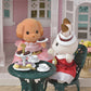 Two Calico Critters figures in elegant outfits enjoy tea and treats at a petite patio table, complete with the Tea and Treats Set featuring teacups, sweets, macarons, and delightful miniature accessories in a charming garden scene.