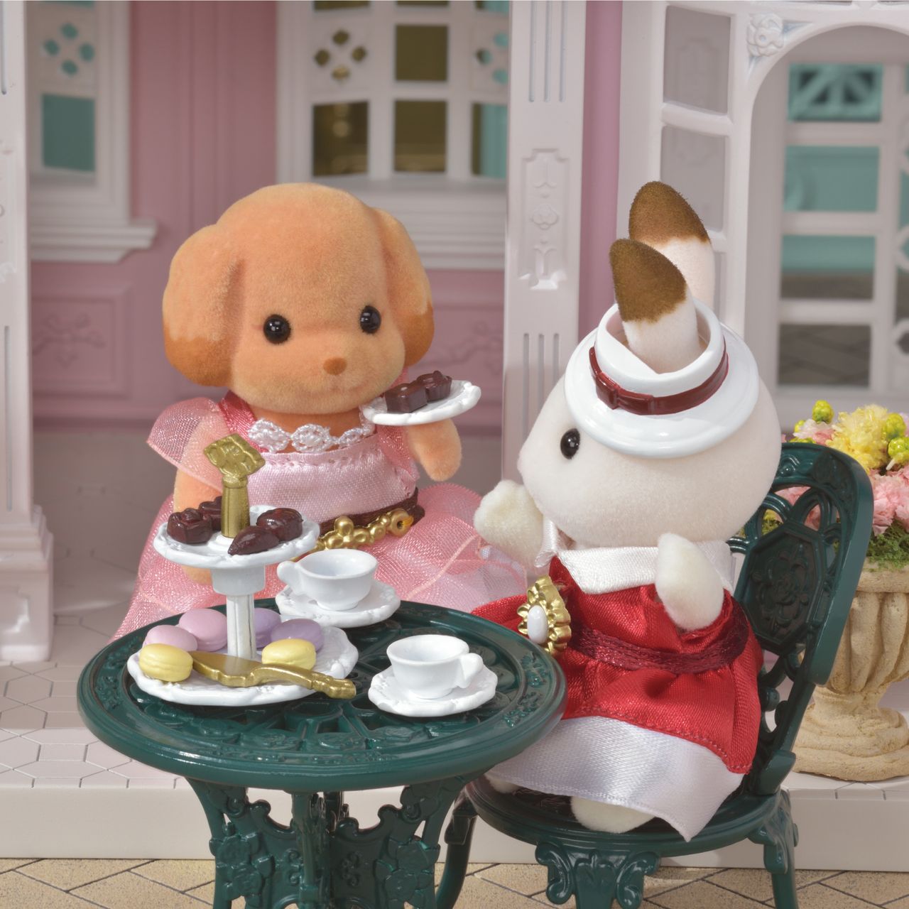 Two Calico Critters figures in elegant outfits enjoy tea and treats at a petite patio table, complete with the Tea and Treats Set featuring teacups, sweets, macarons, and delightful miniature accessories in a charming garden scene.