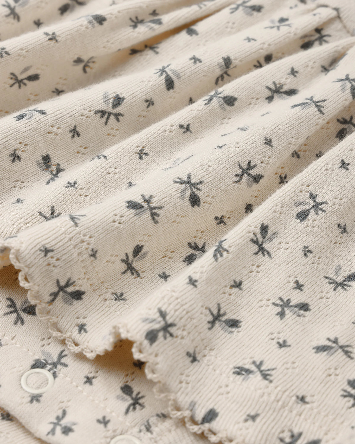 Close-up of a cream-colored fabric from the Quincy Mae Baby Girls’ Pointelle Skirted Bodysuit, featuring small black insect or dragonfly motifs, scalloped edges, a delicate peplum skirt, and snap button closures.