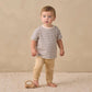 A toddler wearing the Quincy Mae Baby Boys’ and Boys’ Boxy Tee with a relaxed fit and stripes, paired with beige pants, stands on a textured rug beside a small wooden toy car, set against a neutral background.
