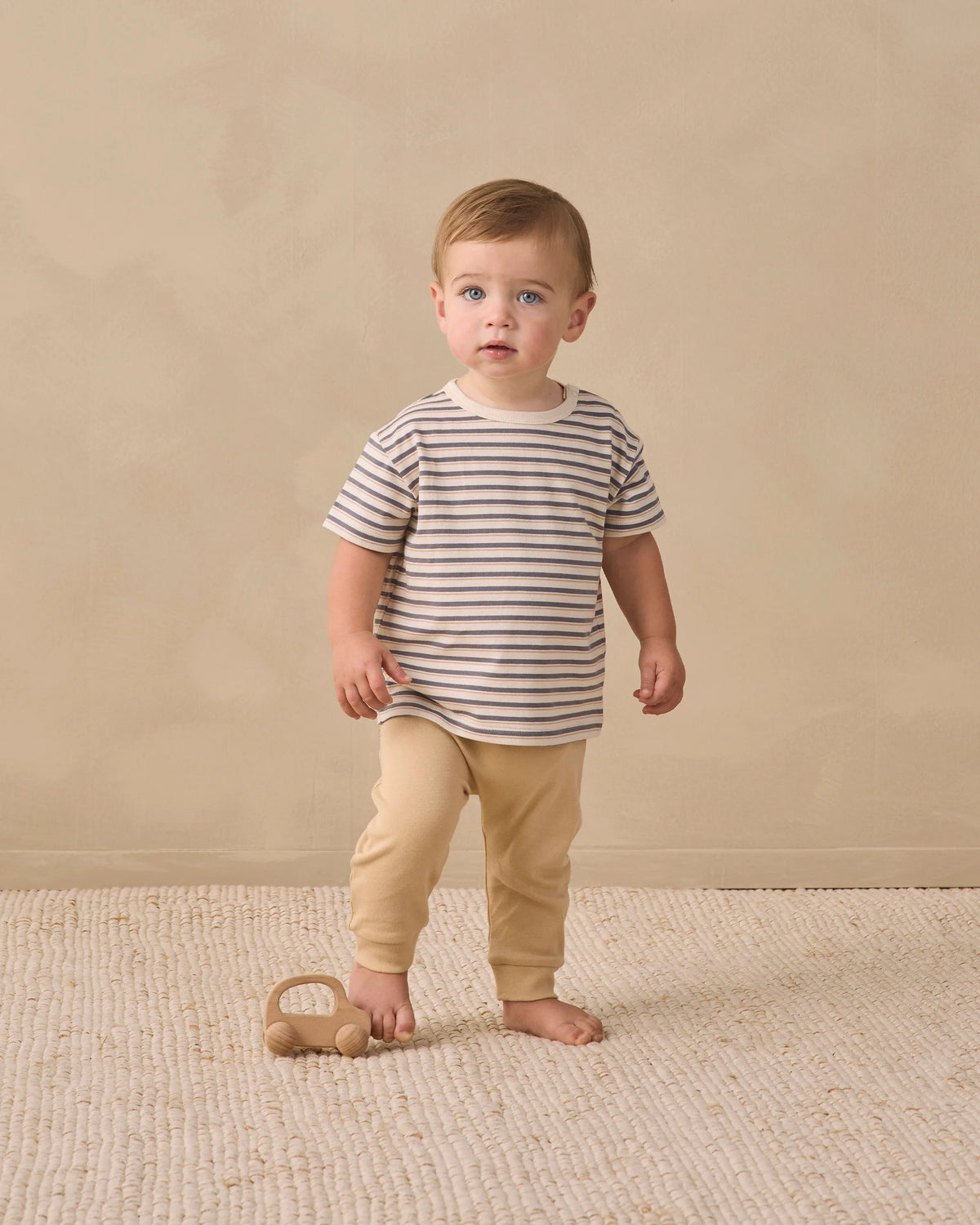 A toddler wearing the Quincy Mae Baby Boys’ and Boys’ Boxy Tee with a relaxed fit and stripes, paired with beige pants, stands on a textured rug beside a small wooden toy car, set against a neutral background.
