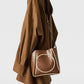 A woman in a brown oversized shirt dress carries the Stella McCartney Logo Raffia Trim Crossbody Bag, crafted from vegan leather and featuring a circular logo with rope detailing.