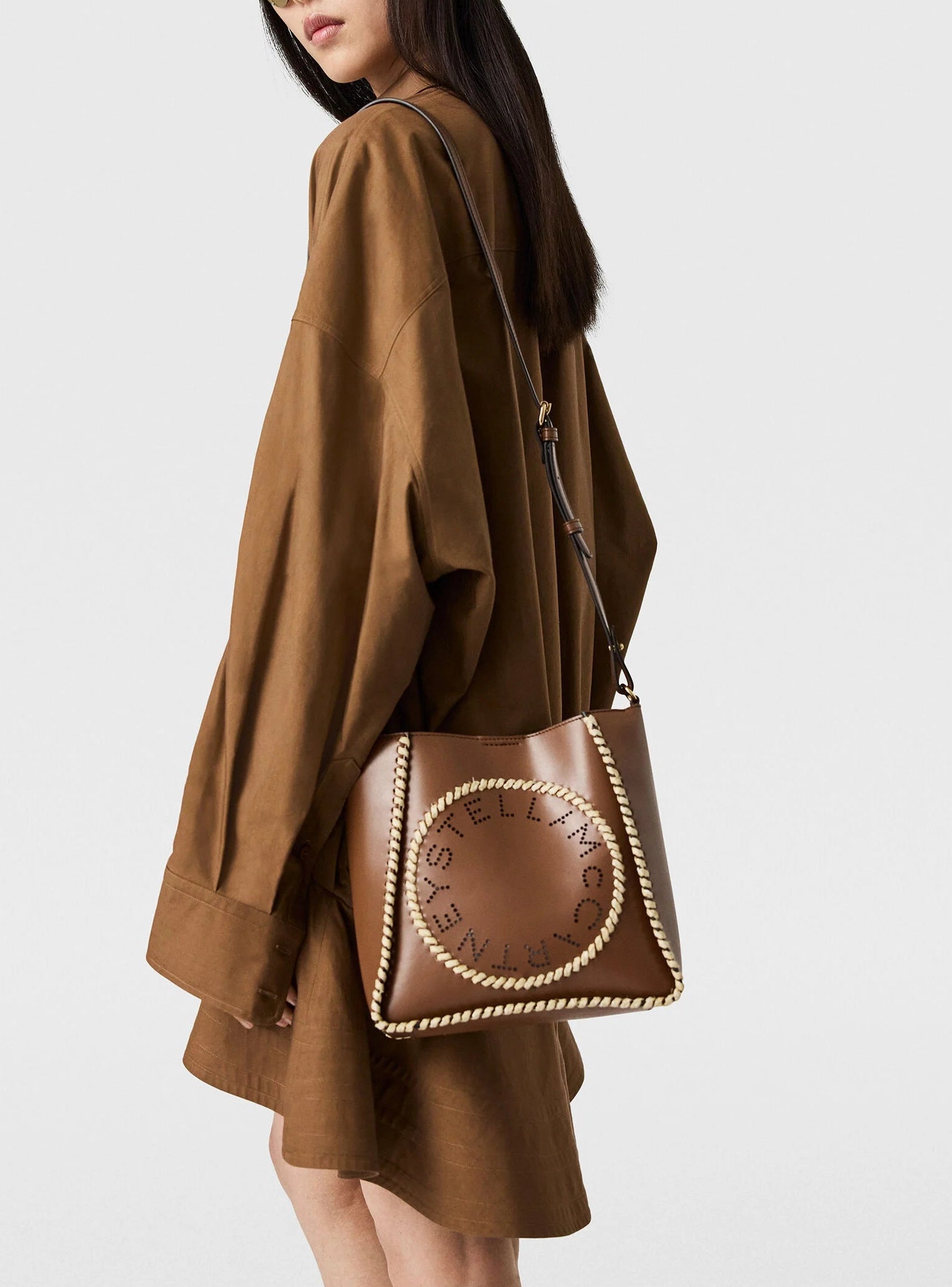 A woman in a brown oversized shirt dress carries the Stella McCartney Logo Raffia Trim Crossbody Bag, crafted from vegan leather and featuring a circular logo with rope detailing.