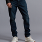 A person wearing Rag & Bone's Fit 3 Authentic Stretch dark blue tapered leg jeans, white sneakers, a striped shirt, and a dark jacket stands against a plain gray background.