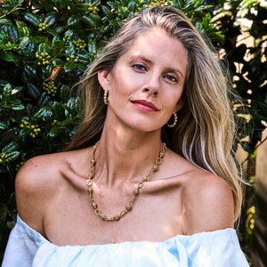 A woman with long blonde hair, wearing a white off-the-shoulder top and Juliska Capucine De Wulf Berry Icon Small Hoop Earrings, poses in front of green foliage.