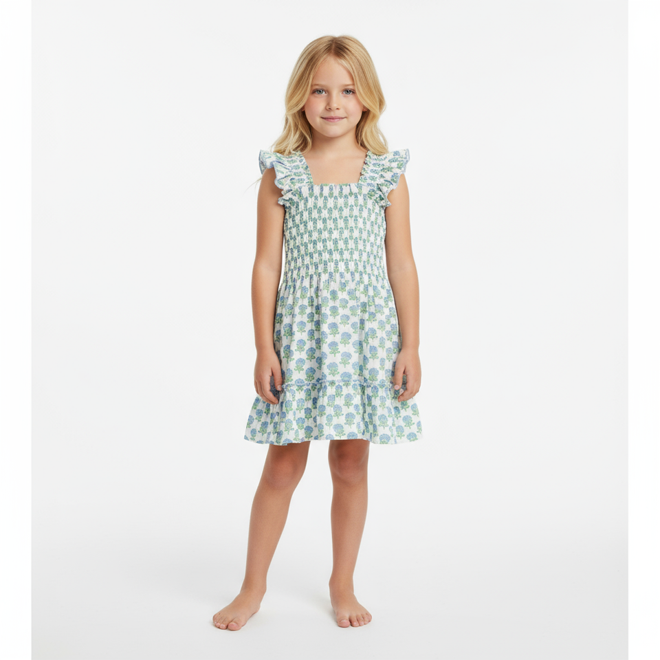 A young child with long blond hair stands barefoot against a plain white background, wearing the Mer Girls' Paloma Dress, which features smocked flutter sleeves and a ruffle twirly skirt.
