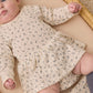 A baby wearing a cream-colored Quincy Mae Baby Girls’ Pointelle Skirted Bodysuit with a delicate floral print and peplum skirt lies on a beige surface, with only the lower half of her face visible.