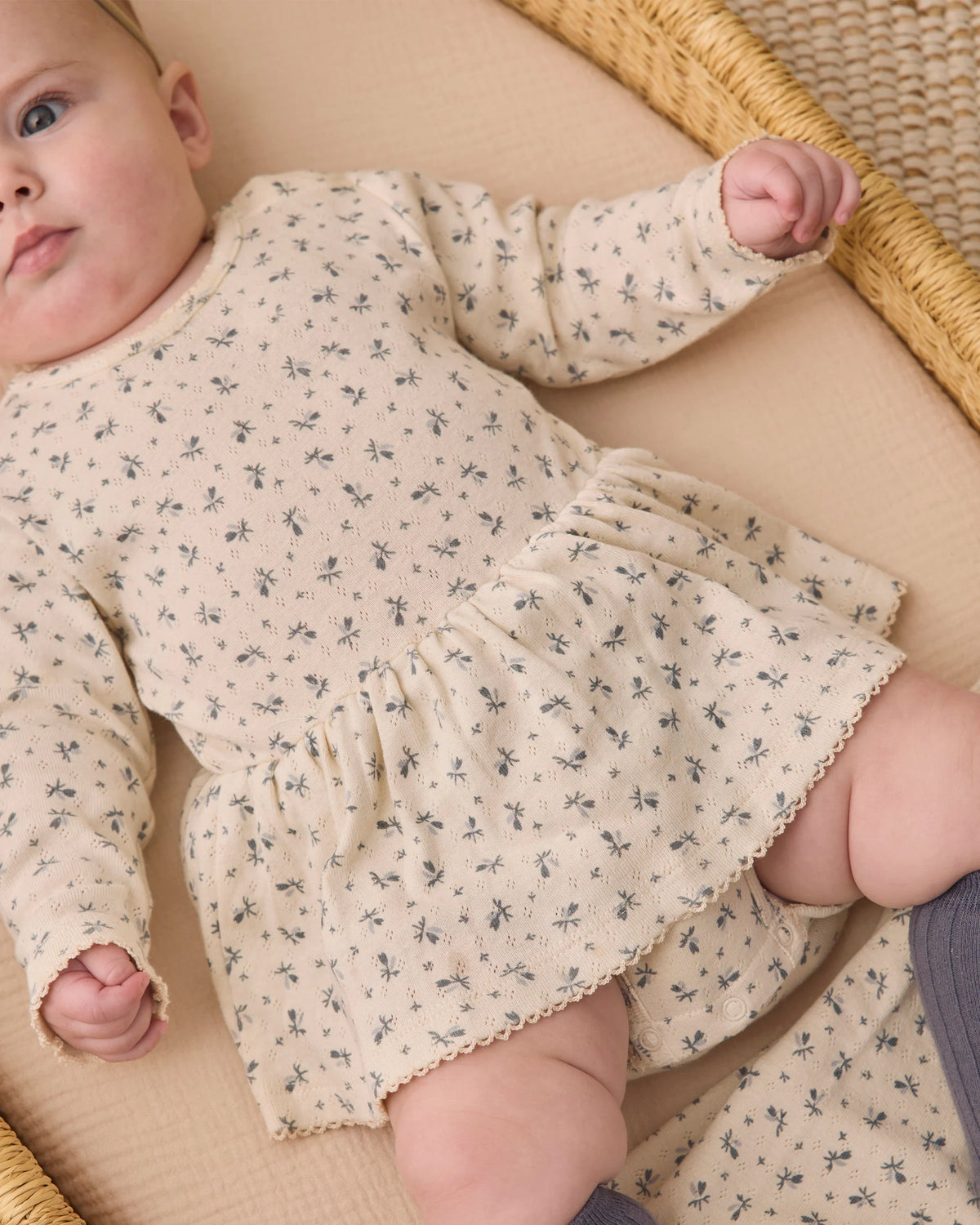 A baby wearing a cream-colored Quincy Mae Baby Girls’ Pointelle Skirted Bodysuit with a delicate floral print and peplum skirt lies on a beige surface, with only the lower half of her face visible.