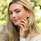 A woman with long blonde hair wears a cream sweater and Juliska Capucine De Wulf Mermaid Garden Petite Stud Earrings, smiling softly outdoors with lush greenery in the background.