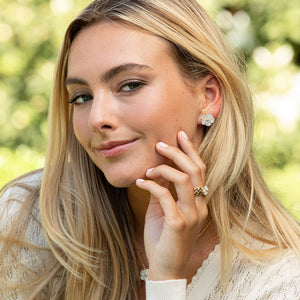 A woman with long blonde hair wears a cream sweater and Juliska Capucine De Wulf Mermaid Garden Petite Stud Earrings, smiling softly outdoors with lush greenery in the background.