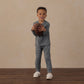 A young boy wearing Rylee & Cru Boys’ Century Tech Jogger in blue stands on a wooden floor, holding a football and smiling at the camera against a beige background.